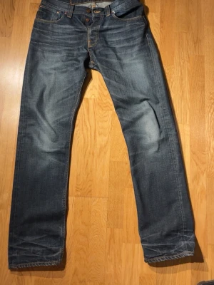 Blå raka jeans från Nudie Jeans W34 L34 - Säljer ett par klassiska blå jeans från Nudie Jeans med raka ben och snygga slitningar. Jeansen har fem fickor, orangea sömmar och är gjorda i slitstark denim. Perfekta för en avslappnad och trendig look.