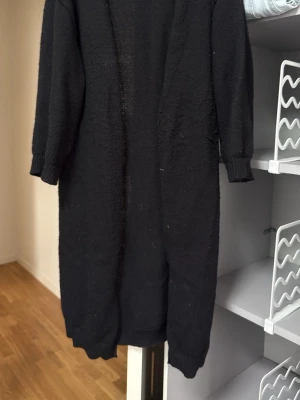 Lång svart stickad kofta - Säljer en lång, svart stickad kofta med ribbade muddar vid ärmslut och nederkant. Koftan har en öppen front, tills under knäna 