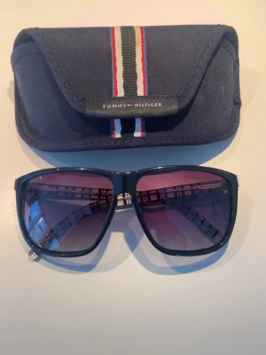 Svarta solglasögon Tommy Hilfiger - Snygga svarta solglasögon från Tommy Hilfiger med fyrkantig form och mörka tonade glas. Bågarna är i plast och kommer med ett blått fodral med Hilfiger-logga och randig detalj i rött, vitt och blått. Perfekta för soliga dagar! Pris kan självklart diskuteras! Allt är självklart äkta! Hör av er vid minsta fundering!
