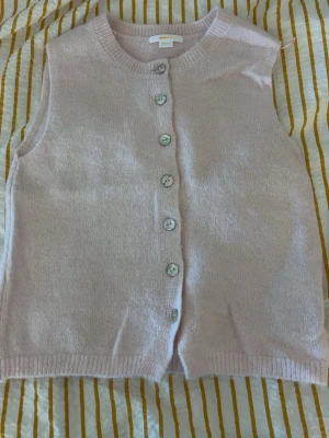 Ljusrosa stickad väst Gina Tricot - Mysig ljusrosa stickad väst från Gina Tricot med rund halsringning och pärlemorknappar framtill. Västen är ärmlös och har ribbade kanter vid hals och nederkant. Perfekt att styla över en t-shirt eller skjorta för en trendig lager-på-lager-look.