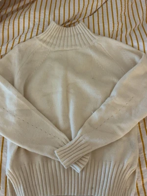 Beige stickad polotröja - Mysig beige stickad polotröja med bred ribbad mudd vid hals, ärmslut och nederkant. Tröjan har lång ärm och är tillverkad i mjukt material som känns skönt mot huden. Perfekt för kyliga dagar och enkel att matcha med olika outfits. Aldrig andvänd!