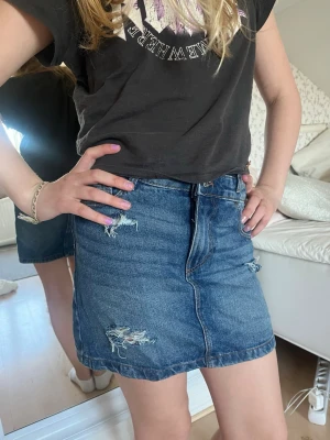 Blå jeanskjol med slitningar - Säljer en klassisk blå jeanskjol i kort modell med slitna detaljer framtill och bakfickor. Kjolen har en rak passform och stängs med knapp och dragkedja framtill. Det är dubbel knappar så man kan justera smått i midjan. Storlek xs💘