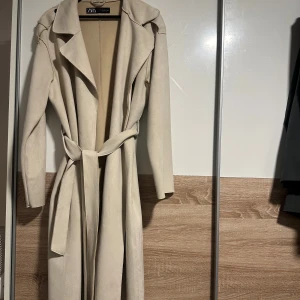 Beige trenchcoat från Zara - Stilren beige trenchcoat från Zara med klassisk krage och knytskärp i midjan. Kappan har en lång, rak passform och är tillverkad i ett mjukt material som ger en lyxig känsla. Perfekt för att lyfta vilken outfit som helst.