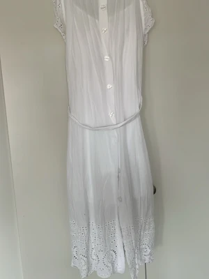 Vit sommarklänning Tommy Hilfiger - Vit knälång klänning från Tommy Hilfiger i bomull med broderade detaljer nertill och knappar framtill. Klänningen har korta ärmar med spets och ett smalt bälte i midjan. Perfekt för varma dagar och har en luftig, feminin look.