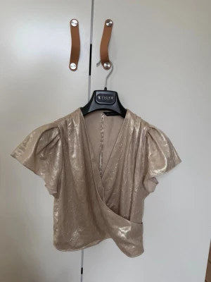 Champagnefärgad omlottopp med puffärm - Snygg champagnefärgad omlottopp från Tiger of Sweden med korta puffärmar och lätt glansigt material. Toppen har v-ringning och ett subtilt mönster i tyget som ger extra lyxig känsla. Perfekt för dig som vill sticka ut lite extra. S/m