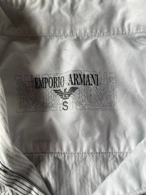 Randig skjorta från Emporio Armani - Snygg vit skjorta från Emporio Armani med tunna svarta ränder och klassisk krage. Skjortan har en bröstficka med lock och broderad logga, samt knappar framtill. Perfekt för dig som gillar stilrena detaljer och exklusiv känsla.