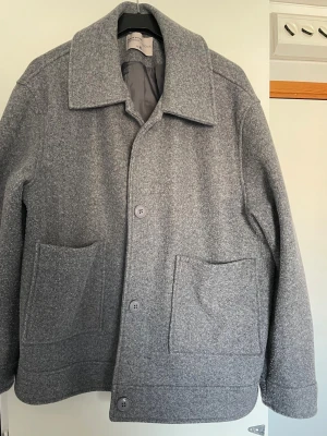 Grå ullkappa  -  grå ullkappa från My wear women med rak passform, stora framfickor och bred krage. Kappan har fyra knappar framtill och är helfodrad i grått tyg. Perfekt för dig som gillar enkel och stilren design.