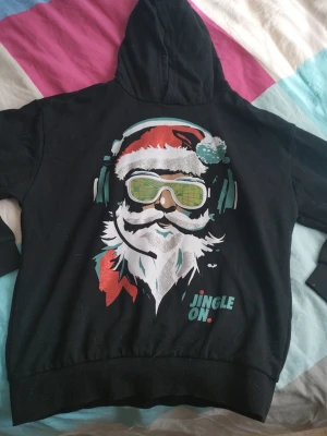 Svart hoodie med julmotiv från F&F - Svart hoodie från F&F med huva och ett färgglatt julmotiv framtill. Trycket visar en figur med tomteluva och hörlurar samt texten 'JINGLE ON.' i turkos och rött. Mjuk bomullskänsla och ribbade muddar vid ärmar och nederkant.