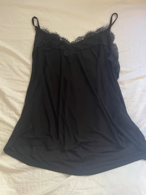 Svart linnetopp med spetsdetaljer - Snygg svart linnetopp med tunna axelband och spetsdetaljer längs urringningen. Toppen har en lös passform och är tillverkad i ett mjukt, följsamt material som ger en elegant känsla. Perfekt att styla lager på lager eller bära som den är.