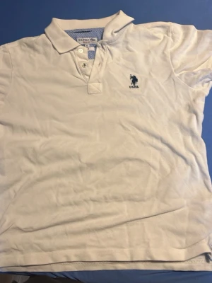 Vit pikétröja från U.S. Polo Assn. - Klassisk vit pikétröja från U.S. Polo Assn. med blå logga broderad på bröstet. Tröjan har krage, knappar framtill och är tillverkad i bomull. Perfekt för en clean och sportig look.