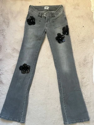 Grå jeans med svarta paljettblommor - Unika grå jeans från 157 med utsvängda ben och stora svarta blommor i paljetter på fickor och ben. Snygg detalj med dubbla knappar i midjan och dekorativa sömmar. Perfekta för dig som vill sticka ut med coola detaljer och trendig look.