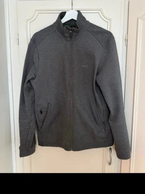 Mörkgrå ziptröja från BOSS - Snygg mörkgrå ziptröja från BOSS med hög krage och diskret logga på bröstet. Tröjan har två sidofickor med dragkedja och är tillverkad i mjukt material som känns bekvämt. Perfekt för en avslappnad och stilren look.