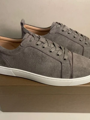 Gråa sneakers från Christian Louboutin - Stilrena gråa sneakers i mocka från Christian Louboutin med klassisk röd sula och vita kanter. Lågt skaft, ton-i-ton snörning och diskret design som passar till det mesta. Kommer med originalkartong och dustbag.