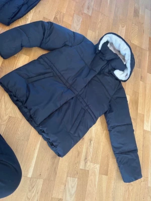 Svart pufferjacka med fuskpäls - Snygg svart pufferjacka med stor huva fodrad i vit fuskpäls. Jackan har en boxig passform och breda kanaler för extra värme. Perfekt för kalla vinterdagar och ger en cool streetwear-look. Använd 2 gånger 