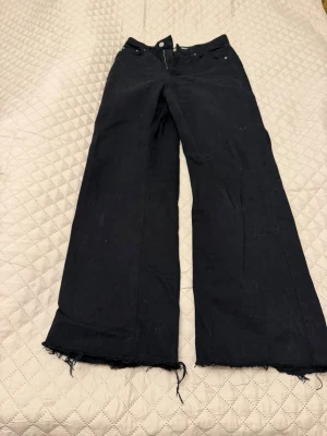 Svarta vida jeans med rå kant - Säljer ett par svarta jeans med breda ben och råa, fransiga benslut. Jeansen har klassisk femficksmodell och hög midja. Perfekta för en avslappnad och trendig look. Materialet är denim och färgen är helsvart.