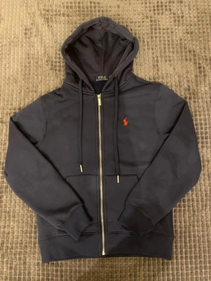 Mörkblå hoodie från Polo Ralph Lauren - Snygg mörkblå hoodie med dragkedja från Polo Ralph Lauren. Klassisk design med huva och dragsnören, samt röd broderad logga på bröstet. Tillverkad i mjukt bomullsmaterial och har fickor framtill. Perfekt för en avslappnad och stilren look.