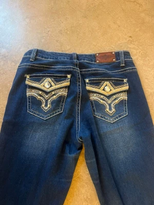Mörkblå jeans med broderade fickor - Snygga mörkblå jeans från Stars & Stripes med unika broderade bakfickor och dekorativa detaljer i guld och silver. Jeansen har kontrastsömmar och en klassisk femficksmodell. Perfekta för dig som vill sticka ut med cool westernstil. Köpte på Sellpy. Knappen sitter lite löst där fram 