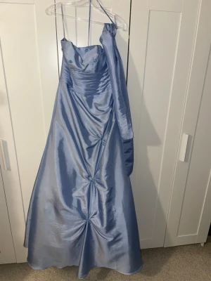 Ljusblå långklänning med pärldetaljer - Elegant ljusblå långklänning i glansigt tyg med halterneck och vackra pärldetaljer framtill. Klänningen har draperingar och dekorativa broderier med pärlor som ger en exklusiv känsla. Tillhörande sjal för att bära över axlarna. Perfekt för bal eller festliga tillfällen.