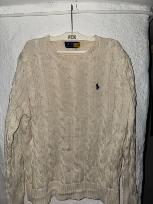 Vit kabelstickad tröja Polo Ralph Lauren - Kabelstickad tröja i offwhite från Polo Ralph Lauren. Klassisk rund halsringning, ribbade muddar och det ikoniska mörkblå broderade logot på bröstet. Tillverkad i mjukt material som ger en skön och stilren look.