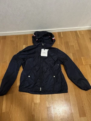 Marinblå vindjacka från Moncler - Snygg marinblå vindjacka från Moncler med huva och dragkedja framtill. Jackan har två fickor med tryckknappar och en vit-röd rand på huvan. Tillverkad i lätt, glansigt material som står emot vind. Perfekt för dig som vill ha en stilren och sportig look.