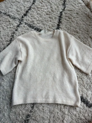 Krämvit stickad tröja med halvärm - Mysig krämvit stickad tröja med rund halsringning och halvärm. Tröjan har en enkel, rak lös passform och är tillverkad i ett mjukt material som känns skönt mot huden. 