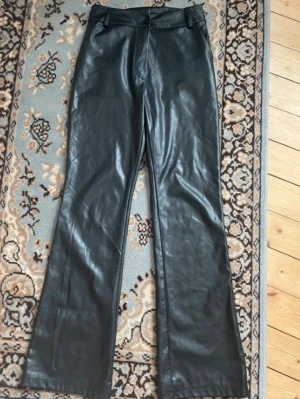 Svarta bootcut byxor i skinnimitation - Säljer ett par svarta bootcut byxor i skinnimitation. Byxorna har hög midja, klassisk knapp och dragkedja framtill samt raka ben med lätt utsvängd form. Perfekta för en edgy och trendig look.