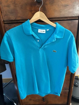 Blå pikétröja från Lacoste - Snygg blå pikétröja från Lacoste med klassisk krage och korta ärmar. Tröjan har den ikoniska krokodilloggan broderad på bröstet och stängs med två knappar vid halsen. Perfekt för en avslappnad men ändå stilren look.