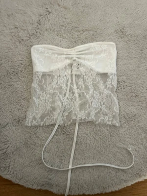 Vit spets tubtopp med snörning - Säljer en superfin vit tubtopp med transparent spets och snörning framtill. Toppen har ett bandeau-snitt och tunna band som kan knytas för extra detalj. Perfekt för dig som vill ha en romantisk och trendig look.