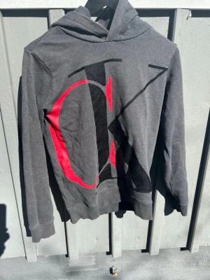 Grå hoodie från Calvin Klein - Snygg grå hoodie från Calvin Klein med stor CK-logga i svart och rött framtill. Klassisk huva och ribbade muddar vid ärmar och nederkant. Perfekt för en avslappnad och trendig look.