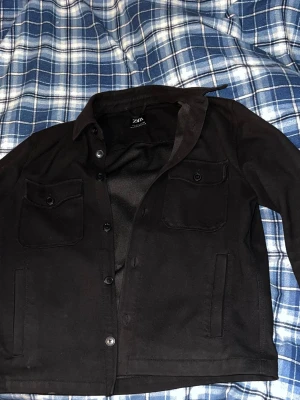 Svart overshirt från Zara - Snygg svart overshirt från Zara med klassisk krage och knäppning framtill. Två bröstfickor med lock och knapp samt två sidofickor. Overshirten har en rak passform och är tillverkad i ett mjukt tyg som känns skönt att bära. Perfekt lager-på-lager-plagg.