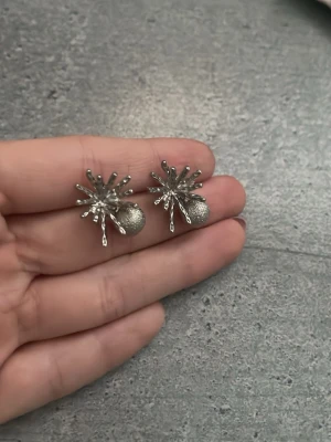 Silvriga örhängen med pärldetalj - Unika silvriga örhängen med en cool stjärnformad design och en rund pärlliknande detalj längst ner. Örhängena är tillverkade i metall och har en modern, edgy look som verkligen sticker ut. Perfekta för dig som vill ha något annorlunda och trendigt.