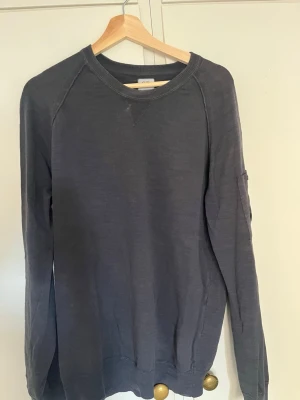 Mörkgrå sweatshirt från C.P. Company - Mörkgrå sweatshirt från C.P. Company med rund halsringning och ribbade muddar. Tröjan har en ficka med den ikoniska linsen på vänster ärm, vilket ger en cool och modern look. Perfekt för dig som gillar streetwear och stilrena plagg.