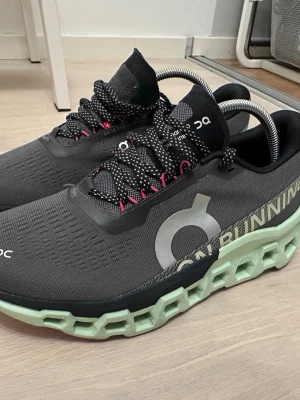 On Running svarta sneakers - Snygga svarta sneakers från On Running med stora vita loggor på sidan och coola detaljer i rosa och mintgrönt. Skorna har en unik, chunky sula i mintgrönt och svarta snören med vita prickar. Ovandelen är i mesh för bra andningsförmåga och sportig look.