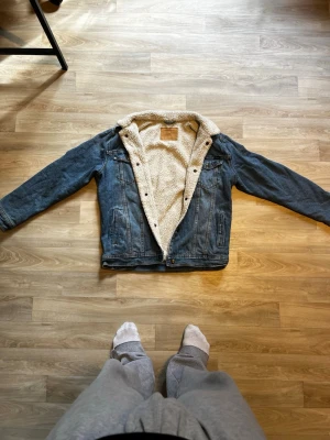 Blå jeansjacka med teddyfoder - Säljer en klassisk blå jeansjacka med vitt teddyfoder och krage. Jackan har två bröstfickor med lock och knappar samt två sidofickor. Perfekt för dig som vill ha en tidlös och snygg look med extra värme. Stängs med knappar framtill.