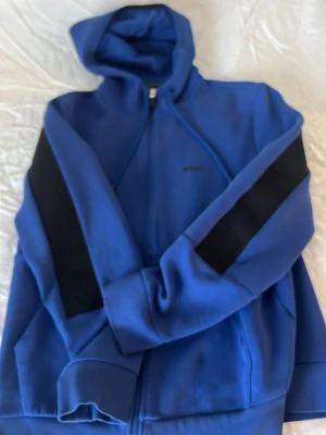 Blå hoodie från BOSS med svarta detaljer - Snygg blå hoodie från BOSS med dragkedja och huva. Tröjan har svarta ränder längs ärmarna och ribbade muddar. Tillverkad i mjukt material som känns skönt mot huden. Perfekt för en avslappnad och trendig stil.