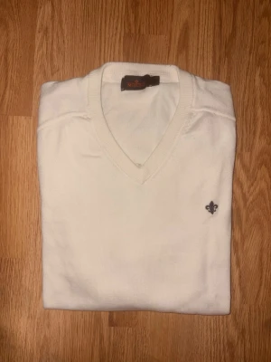 Vit Merino Ull V-ringad tröja från Morris - Klassisk vit V-ringad tröja från Morris med diskret broderad logga på bröstet. Tröjan har en stilren design och är tillverkad i mjukt material som passar perfekt till olika outfits. Enkel att matcha och ett måste i garderoben. Det är Merino Ull.