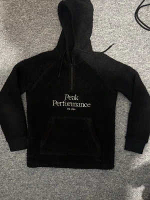  Peak Performance hoodie fleece - Mysig svart hoodie från Peak Performance i teddyfleece med broderad logga på bröstet. Modellen har huva med dragsko, halv dragkedja och stor magficka framtill. Perfekt för dig som gillar en avslappnad och sportig stil.
