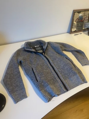 Grå stickad tröja med fleecefoder - Mysig grå stickad tröja med hel dragkedja och två fickor framtill. Insidan är fodrad med mjuk fleece i rutigt mönster i svart, vitt och brunt. Perfekt för kyliga dagar och har en hög krage för extra värme. Storlek M men liten i storleken. Helt ny aldrig använd. 