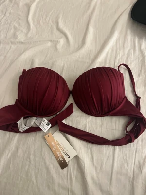 Vinröd bikiniöverdel från Calzedonia - Snygg vinröd bikiniöverdel från Calzedonia i glansig satin med vadderade kupor och fina veckade detaljer. Axelbanden är justerbara och knytband finns i ryggen för perfekt passform. Perfekt för strandhäng eller poolparty!