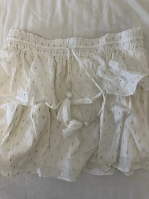 Vit volangkjol med tofsar - Supersöt vit kjol med volanger och små guldfärgade detaljer. Kjolen har resår i midjan och dekorativa tofsar framtill. Tillverkad i ett lätt och luftigt tyg som passar perfekt till sommaren. Ger en bohemisk och avslappnad vibe.