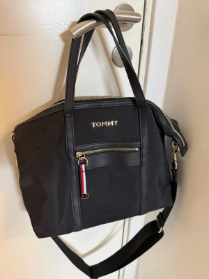 Svart handväska från Tommy Hilfiger - Snygg svart handväska från Tommy Hilfiger med guldfärgade detaljer. Väskan har både handtag och justerbar axelrem, samt en dragkedjeficka framtill med klassisk Tommy Hilfiger-detalj. Perfekt för dig som vill ha en stilren och praktisk väska.