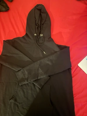 Svart hoodie med ficka och huva - En klassisk svart hoodie med stor känguruficka framtill och justerbar huva med snören. Hoodien har långa ärmar och ribbade muddar. Perfekt för en avslappnad och stilren look. Materialet känns mjukt och bekvämt, troligen bomull eller bomullsblandning.