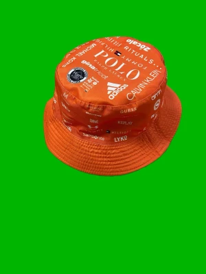 Orange bucket hat med loggor - Cool orange bucket hat med massor av vita loggor från olika märken som Adidas, Calvin Klein och Polo. Hatten har en klassisk form med bred kant och är tillverkad i ett lätt tygmaterial. Perfekt för dig som vill sticka ut och visa din stil.