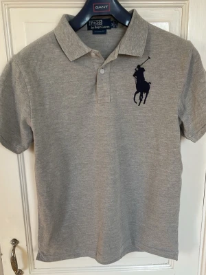 Grå pikétröja Polo Ralph Lauren - Klassisk grå pikétröja från Polo Ralph Lauren med stor mörkblå logga på bröstet och en broderad siffra 3 på ärmen. Tröjan har korta ärmar, krage och knappar framtill. Perfekt för en avslappnad men ändå stilren look