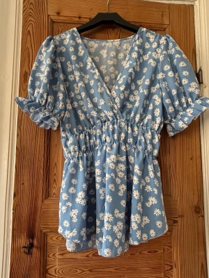 Blommig blå omlottopp med puffärm - Supersöt blå topp med vita blommor och omlott-skärning. Puffiga korta ärmar med volangdetalj och resår i midjan för en smickrande passform. Perfekt för vår och sommar, ger en romantisk och trendig vibe.