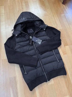 Leon Marcel Hybridjacka – Midnight Navy | Avtagbar Luva | Ny Med Prislapp - Ny med prislapp — Leon Marcels hybridjacka i djup midnattsblå med quiltad pufferkropp och stickade ärmar. Avtagbar luva och lyxiga microfleecefickor som håller händerna varma — detaljer som gör skillnad. Tidlös Skandinavisk design som passar till allt. Perfekt för höst/vinter och kyliga vårdagar. Säljer direkt för ett bättre pris än butik. 📦 Spårad frakt, väl förpackad.