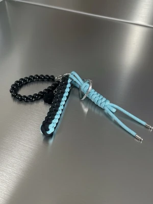Handgjord Paracord Nyckelring / Väskcharm - Unik nyckelring med flätad paracord i ljusblått och svart samt en ring av svarta pärlor. Metallringar och detaljer i silverfärg ger en cool kontrast. Perfekt accessoar att hänga på väskan eller nycklarna för en personlig touch.
