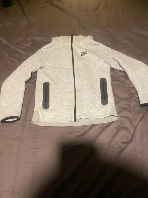 Grå hoodie från Nike med dragkedja - Snygg ljusgrå hoodie från Nike med hel dragkedja framtill och två svarta dragkedjeförsedda fickor. Klassisk Nike-logga på bröstet och huva. Tillverkad i mjukt material som passar perfekt till chill eller träning.