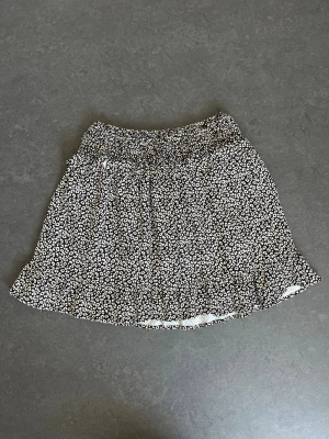 kortkjol med volang - Supersöt svart kortkjol med små vita blommor och volangkant. Kjolen har resår i midjan för skön passform och är perfekt för varma dagar. Lätt och luftig, passar till både sneakers och sandaler.