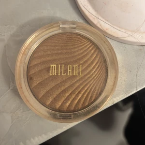 Milani guld highlighter med vågmönster - Säljer en highlighter från Milani i en intensiv guldig nyans. Pudret har ett snyggt vågmönster och kommer i en rund, genomskinlig plastförpackning med guldiga detaljer. Perfekt för att ge glow till ansiktet och framhäva kindbenen.